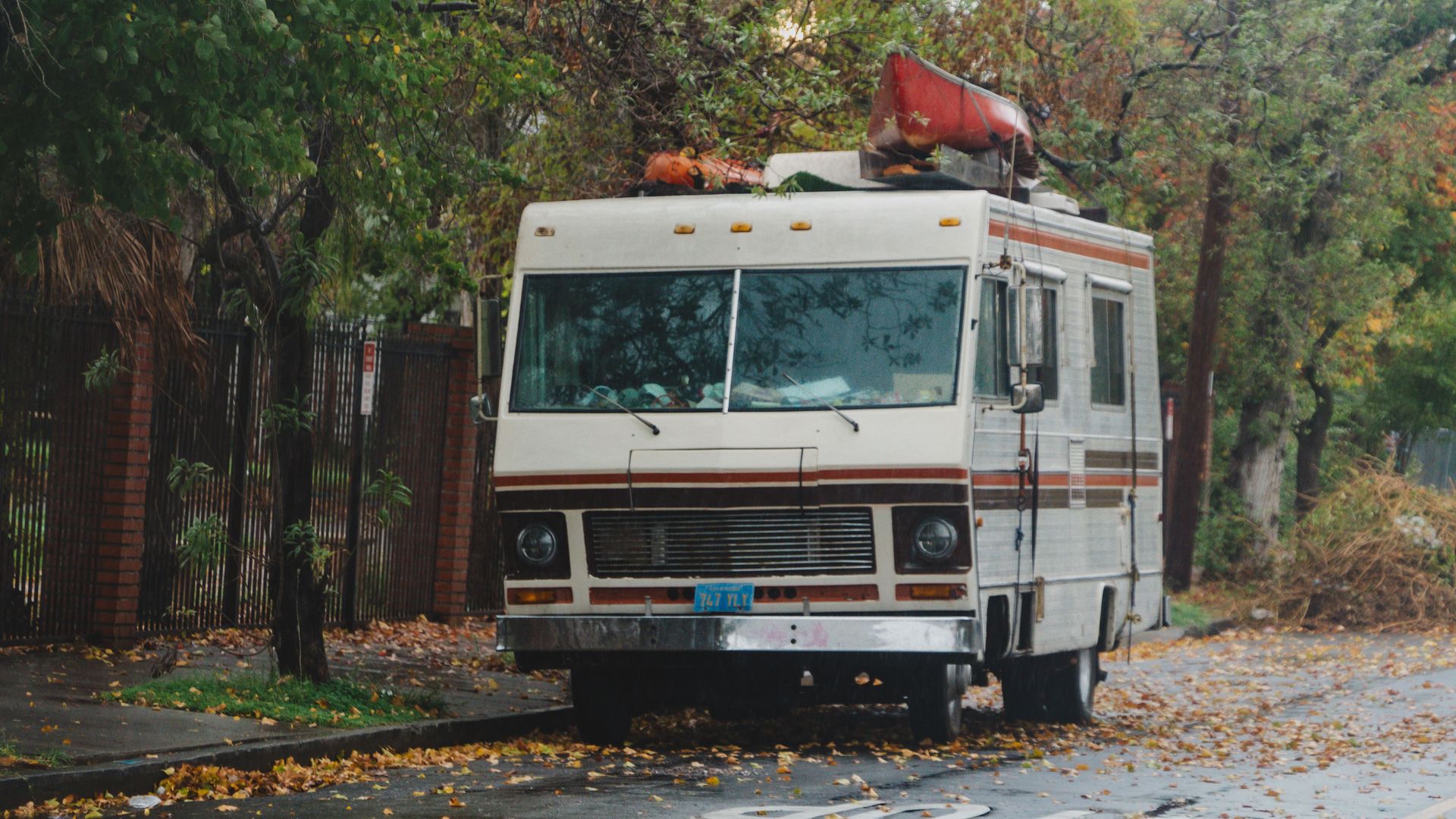 A vintage RV with a red canoe on top parked on a tree-lined street in an autumn setting.