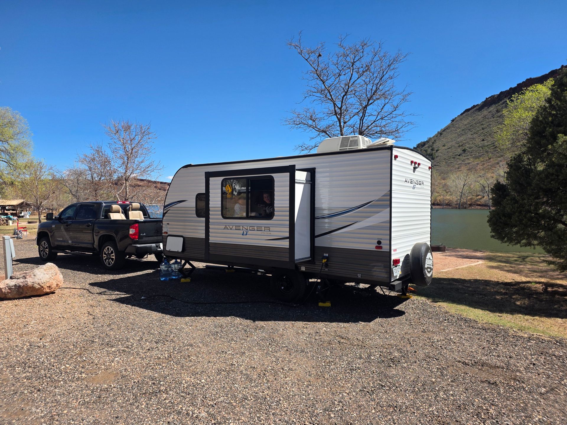 A scenic daytime camping setup with a caravan and truck by a lake in a natural setting.
